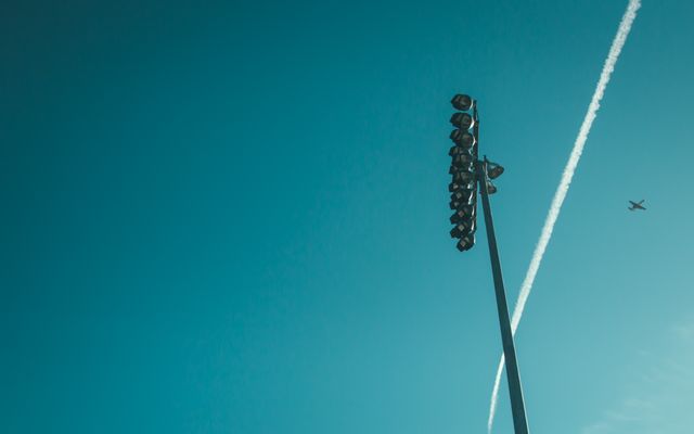 Airplane flying in a clear blue sky next to a tall stadium light post. A vapor trail follows the plane, highlighting the vastness and the simplicity of the open sky. Ideal for themes involving travel, aviation, minimalism, and future possibilities.