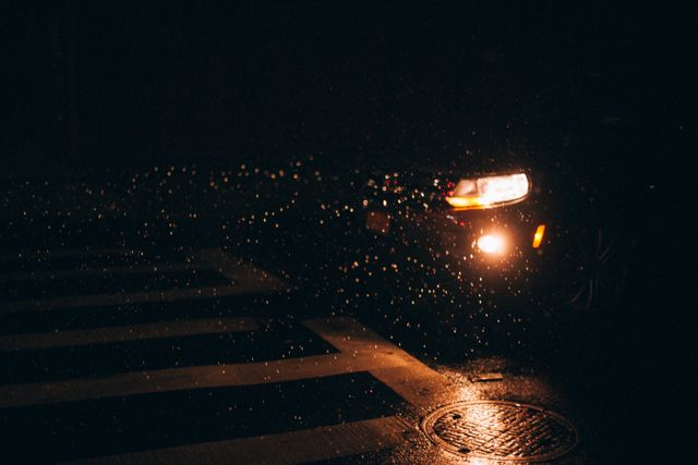 Car driving at night under rainy conditions with headlights on. The wet road reflects the light creating a dramatic and moody atmosphere. Useful for themes related to driving, automobile safety, night scenes, and urban life.