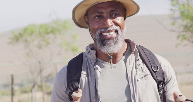 Happy older man smiling while hiking in nature, carrying a backpack and wearing a hat. This image can be useful for promoting outdoor activities, active lifestyles, travel, adventure tourism, and senior fitness programs.