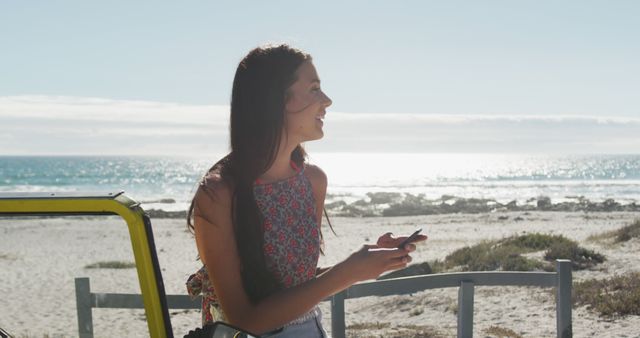 A young woman is standing near a beach, holding a mobile phone and smiling, enjoying a sunny day. The background shows a clear sky and sea. Perfect for travel, vacation, lifestyle, and technology-related projects.