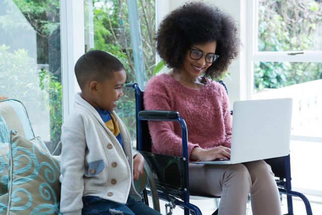 Mother in wheelchair using laptop with her son in a bright living room. They are smiling and enjoying their time together. This image can be used for themes related to family bonding, technology use, disability awareness, and home life.