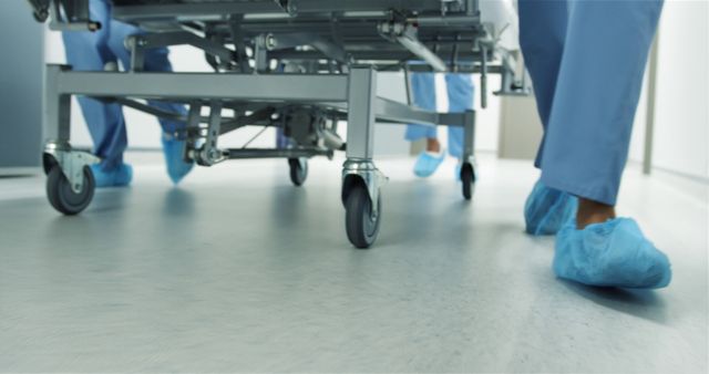 Image depicts lower legs of medical professionals in blue scrubs wheeling a hospital bed down a corridor. Ideal for healthcare articles, patient care brochures, medical training materials, and healthcare facility promotions.