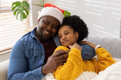African american couple cuddling on sofa wearing santa hat and cozy knit sweater holiday