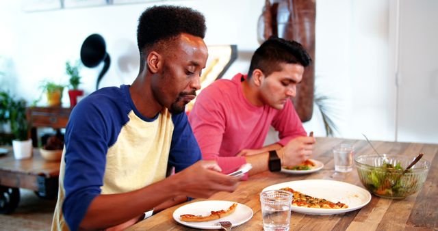 Two friends enjoying pizza while checking their mobile phones at a dining table. Casual setting reflects modern lifestyle and habit of using technology during meals. Suitable for themes related to social interactions, friendship, contemporary routines, and digital habits.