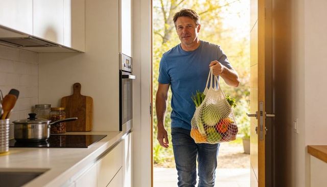 Middle-aged man carrying mesh bag of fresh produce entering sunlit modern kitchen