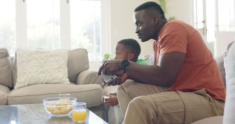 African American father and son playing video games on sofa, bonding during relaxed home gaming