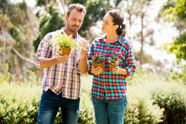 Young couple standing in a lush garden, each holding potted plants. Both are dressed in casual plaid shirts and jeans, engaging in a gardening activity. This image is perfect for use in lifestyle blogs, gardening websites, or advertisements promoting outdoor activities and hobbies.