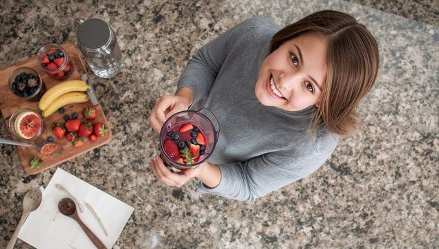 Smiling teen holding blender cup preparing vibrant berry smoothie on granite countertop