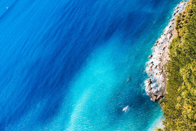Depicts a stunning aerial view of the clear blue waters of the sea meeting a rocky shoreline. Ideal for use in travel and tourism advertisements, nature and vacation brochures, and scenic landscape articles.