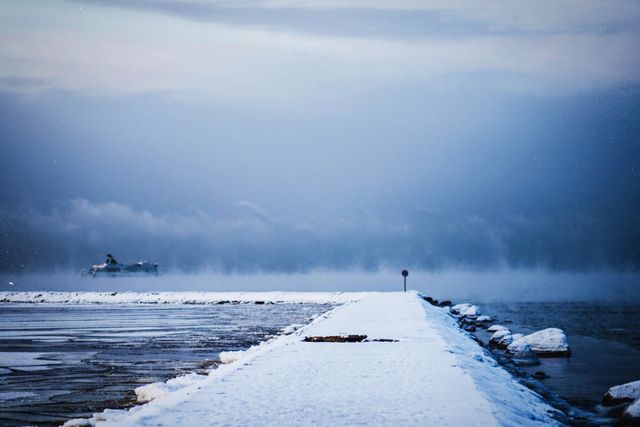 Foggy winter scene with a lone ship in the distance and a snow-covered pier stretching into the sea. Useful for illustrating wintertime solitude, maritime travel, serene nature, and cold weather themes. Ideal for use in travel guides, blogs, and environmental awareness campaigns.