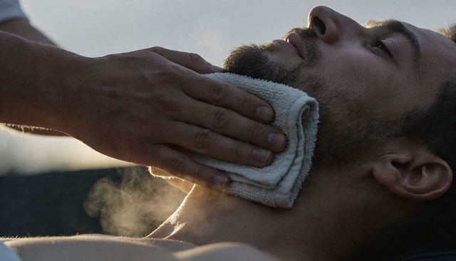 Barber applying warm towel to beard and neck at sunrise for relaxed grooming, steam therapy