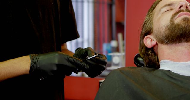 A bearded man relaxing with his eyes closed while receiving a haircut at a barbershop. The barber, wearing black gloves, is preparing to use a pair of scissors. Useful for promoting barbershops, grooming products, men’s fashion, self-care services, and hairstyling tutorials.