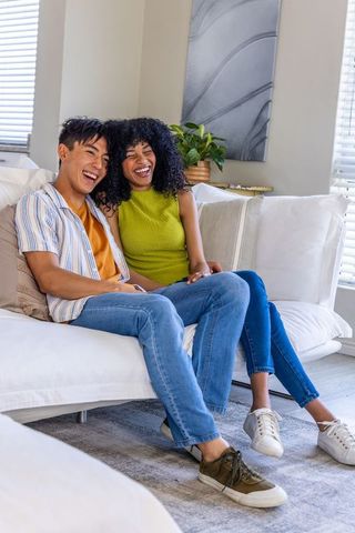 Diverse couple laughing and relaxing on white sofa in bright cozy minimalist living room