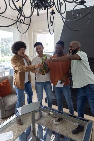Diverse men celebrating and clinking beer bottles at home around glass coffee table with laptop