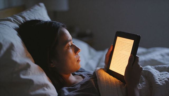 Woman reading backlit e-reader in bed at night under quilted blanket