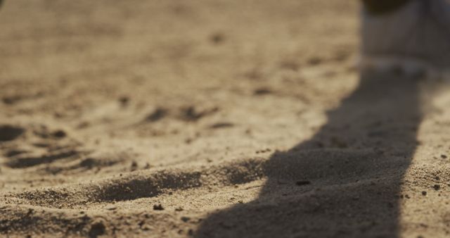 Runner's shadow is cast on a sandy surface under the warm light of sunset, highlighting the texture of the sand. Useful for themes such as sports, outdoor activities, running, fitness, and natural settings. Can be used in promotions for sports equipment, outdoor events, or fitness lifestyle imagery.