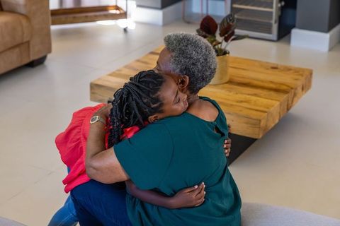 Senior african american woman hugging young woman on ottoman in modern sunlit living room