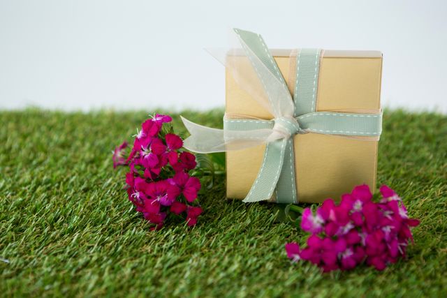 Gift box with ribbon placed on grass with vibrant flowers. Ideal for use in themes related to celebrations, nature, gifts, and outdoor events. Perfect for greeting cards, invitations, and promotional materials for special occasions.