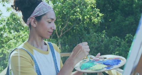 Woman painting outdoors with colorful palette and brush wearing denim overalls headscarf