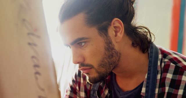 Young male artist focuses intently, creating artwork on canvas in studio. He's wearing a plaid shirt, and the light streams in from the side, emphasizing his dedicated concentration. Suitable for themes related to artistic process, creativity, inspiration, hard work, and passion for art.