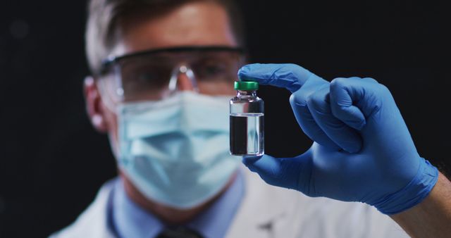 Scientist in lab coat holding a vaccine vial and wearing safety goggles, a mask, and blue gloves. Great for medical or health-related articles, research publications, or pharmaceutical promotions.