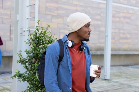 African American man standing in urban plaza holding takeaway coffee wearing beanie and headphones