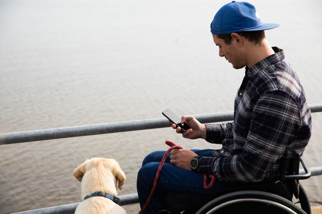 Side view of a young Caucasian man in a wheelchair taking a walk with his dog in the countryside, by the sea, using smartphone