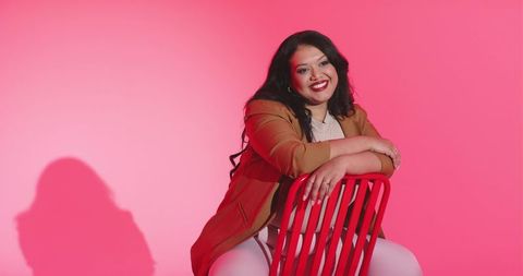 Confident mid-adult woman sitting backward on red chair smiling in tan blazer