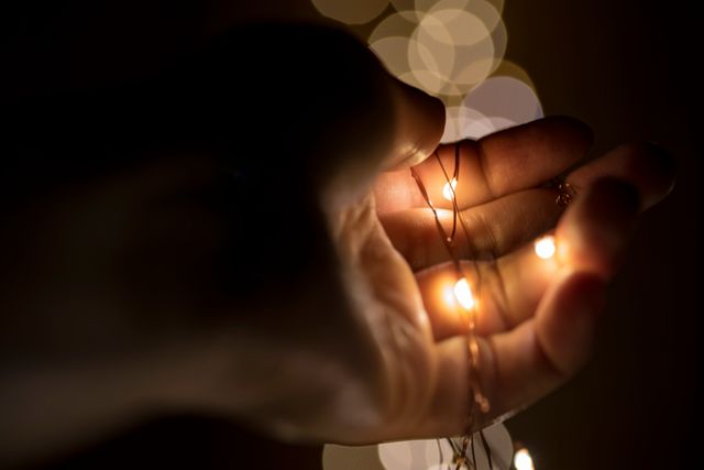 Hand holding warm glowing string lights in a dark setting, creating a festive mood. Suitable for holiday season themes, celebration visuals, cozy ambiance, and light-related concepts.