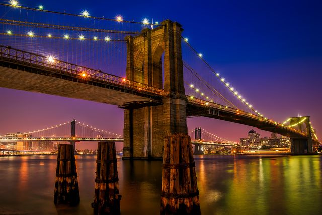 Brooklyn Bridge illuminated at night with city skyline in the background and calm water reflecting lights. Ideal for travel promotions, cityscape posters, greeting cards, tourism brochures.