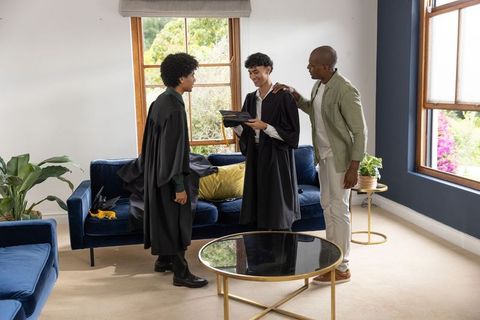 Two young graduates celebrating with family in modern living room wearing caps and gowns