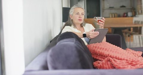 Senior woman relaxing on sofa with tablet and cup of tea, cozy living room scene