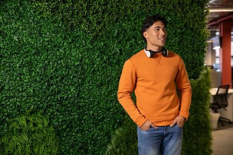 Young asian man standing smiling by green living wall in modern office wearing headphones