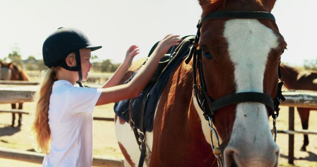Young girl participating in a summer riding camp seen placing a saddle on a horse. She is wearing a protective helmet, indicating a focus on safety and equestrian training. Useful for themes related to outdoor activities for children, summer camps, pets and animal care.