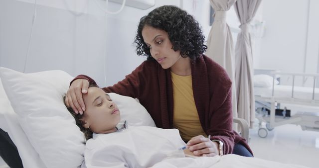 Mother gently placing hand on child's forehead while child rests in hospital bed. Image useful for healthcare, family support, pediatric care, medical treatment, and hospital-related content. Suggests themes of care, nurturing, and the emotional bond between parent and child during illness.