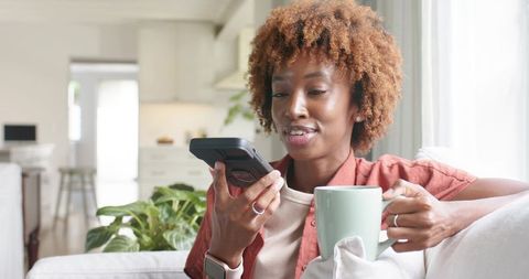 African american woman using smartphone voice assistant while holding coffee mug on couch