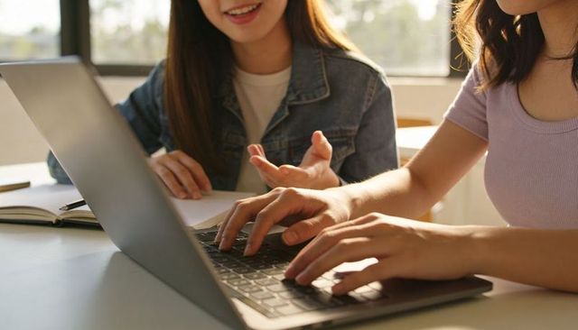 Two Asian Students Collaborating on Laptop and Notebook at Study Table in Sunlit Campus