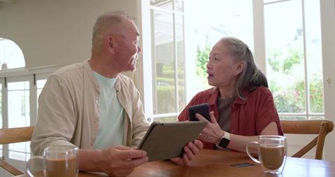 Senior asian couple sharing tablet and smartphone at sunlit dining table