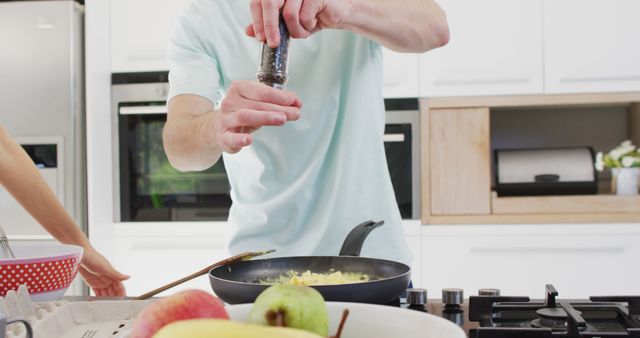 Foreground focuses on man adding seasoning to a frying pan on modern kitchen stove. Captured hands in motion, indicative of culinary action, home cooking or quick meal prep content. Suitable for articles, blogs, advertisements promoting cooking, kitchenware products, culinary skills, or healthy home-cooked meals. Background equipment on countertop suggests a contemporary and organized kitchen environment.