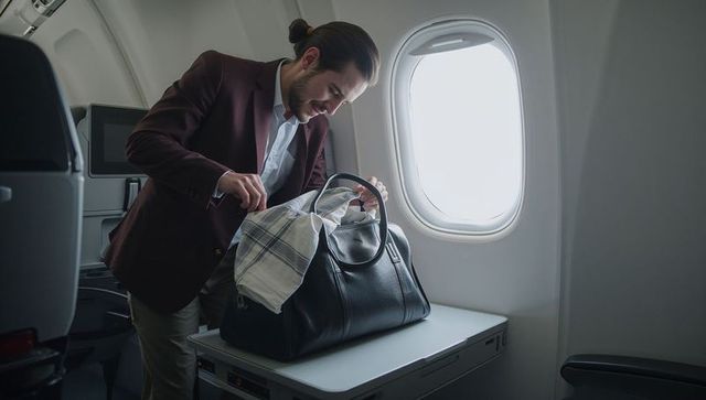 Business traveler searching leather duffel at airplane window seat, arranging scarf