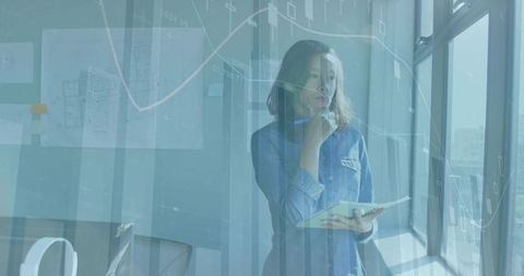 Contemplating businesswoman holding notebook by window with financial graphs overlay