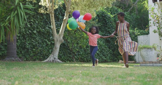 Happy african american mother and daughter holding balloons and running in garden. Lifestyle, domestic life, family, and togetherness.