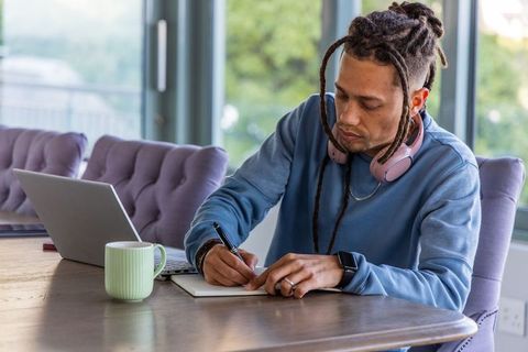 Focused man writing in notebook with laptop and headphones at sunlit home workspace