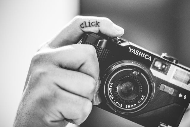 Close-up photo of a hand holding a vintage Yashica camera, focusing on the click tattoo on the index finger. This image is in black and white, emphasizing the nostalgic and retro feel of the scene. Best suited for themes related to photography, creativity, and vintage equipment. Ideal for use in articles, blogs, websites, and marketing materials that discuss photography, retro tech, and creative professions.