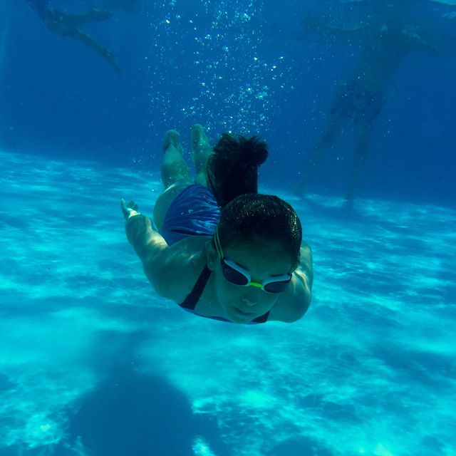Woman swimming underwater in clear blue pool while wearing goggles. Ideal for use in content related to swimming, aquatic activities, fitness, healthy lifestyle, summer, relaxation, and leisure. Suitable for blogs, articles, advertisements, and promotional material focusing on outdoor exercise and recreational activities.