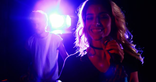 A young Caucasian woman is singing into a microphone, with a young Caucasian man in the background, a DJ, in a setting with vibrant stage lighting, with copy space. Their performance exudes energy and excitement, capturing the essence of live music entertainment.