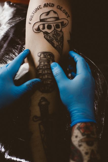 Professional tattoo artist tattooing arm with detailed design of skull wearing bandana and hat. Artist wears protective blue gloves, emphasizing hygiene. Ideal for use in articles about tattoo art, studios, or professional tattoo artists.