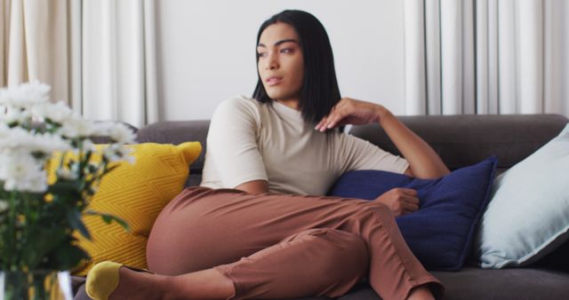 A woman is lounging comfortably on a couch with colorful cushions and floral decorations, dressed in a beige top and brown pants. Her thoughtful expression suggests a moment of introspection and serenity. Ideal for use in lifestyle blogs, home decor magazines, promoting relaxation products, or advertisements for modern living spaces.