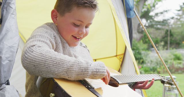 Happy caucasian boy sitting in tent in garden and playing guitar. spending time alone at home.