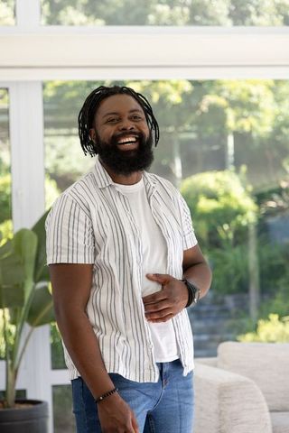 African american man laughing in sunroom wearing striped shirt and jeans, casual lifestyle